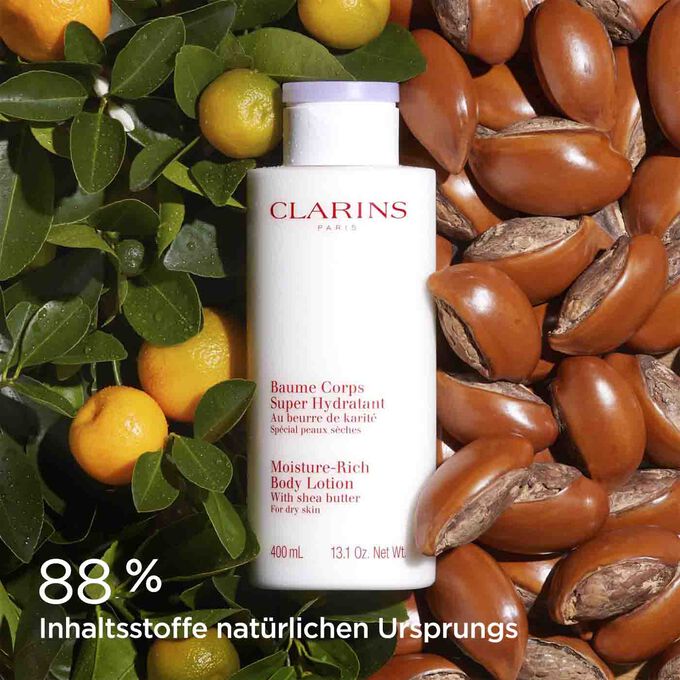 Clarins Baume Corps Body Lotion, umgeben von Shea-Nüssen und Orangen, um die natürlichen Inhaltsstoffe zu verdeutlichen.