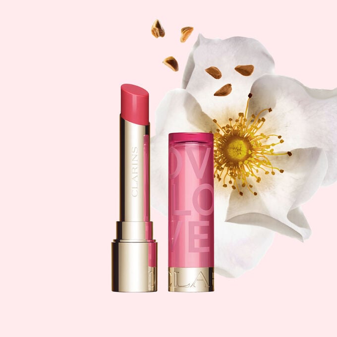 Packshot des Lip Balm aus der Love Collection in limitierter Edition vor einer Rose, die sich wiederum vor einem hellrosa Hintergrund befindet, wodurch die lebendige Farbe des Lip Balm herausgestellt wird.