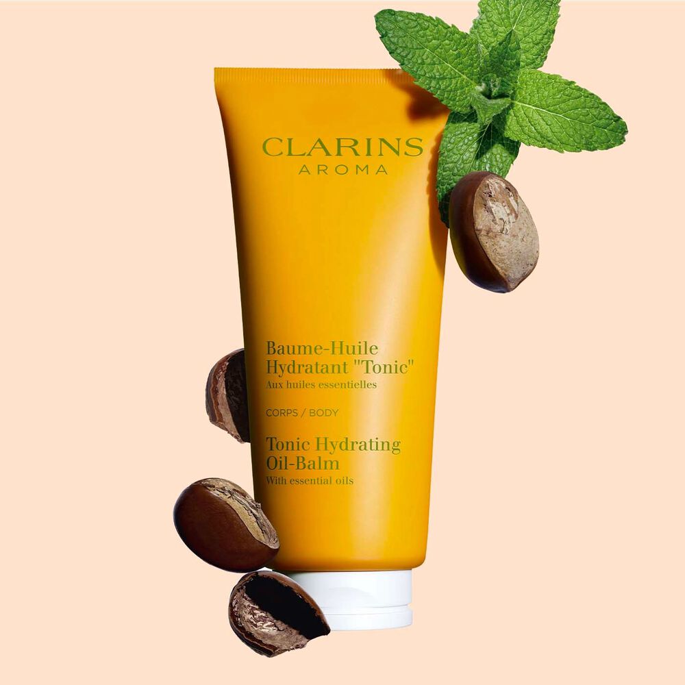 Tube Clarins Baume-Huile Hydratant Tonic, neben einigen Minzblättern und Sheanüssen auf pfirsichfarbenenm Hintergrund.
