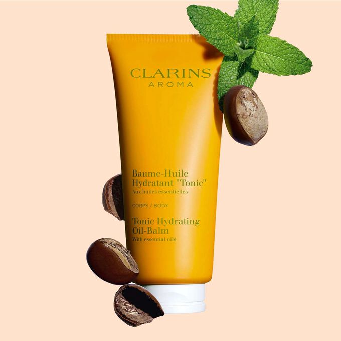 Tube Clarins Baume-Huile Hydratant Tonic, neben einigen Minzblättern und Sheanüssen auf pfirsichfarbenenm Hintergrund.
