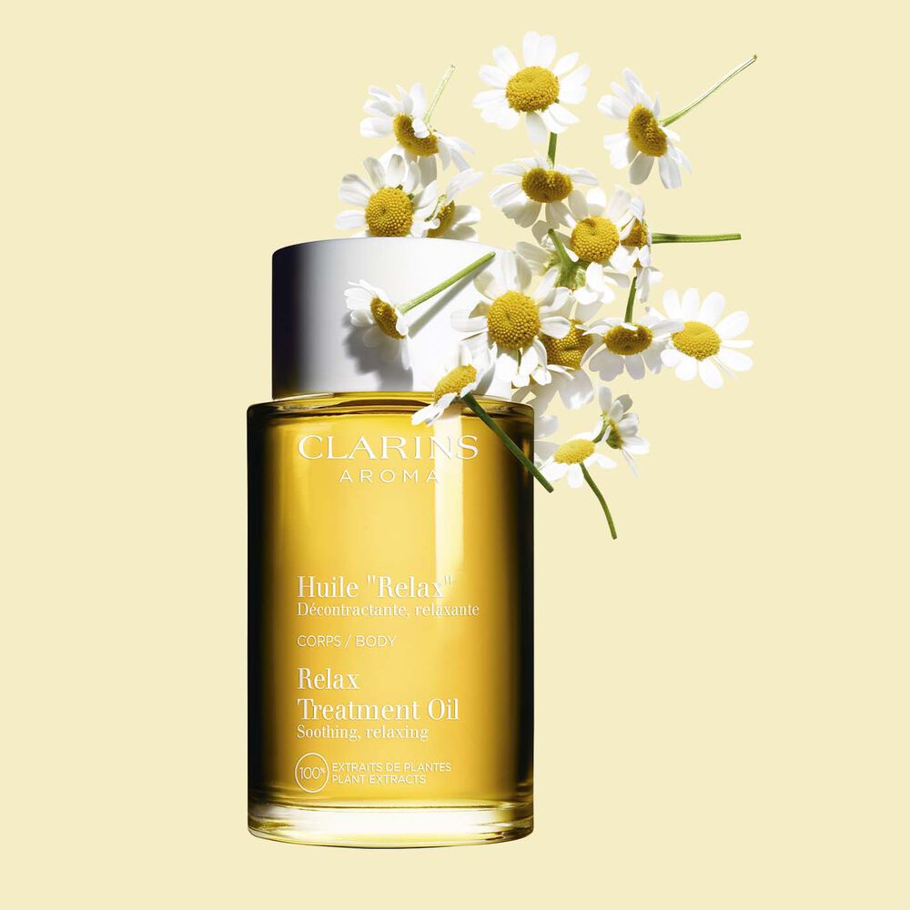 Glasflakon Clarins Körperpflegeöl Huile Relax mit Kamillenblüten auf einem blassgelben Hintergrund